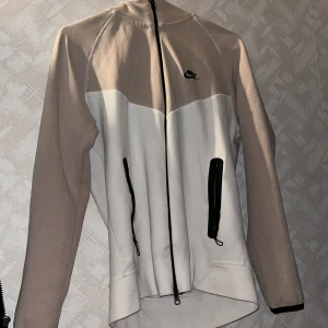 Nike beige och vit vindjacka - Nike vindjacka i beige och vitt med svarta dragkedjor och logga på bröstet. Jackan har lång ärm, hög krage och två praktiska fickor framtill. Perfekt för dig som gillar sportig och clean stil.