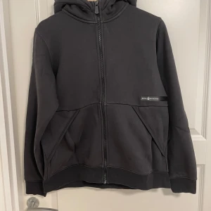 Svart hoodie från Sail Racing - Svart hoodie från Sail Racing med dragkedja hela vägen, huva och två stora fickor framtill. Mjuk insida och diskret logga på bröstet. Perfekt för dig som gillar en clean och stilren look. Passar dig som vill ha en bekväm och snygg tröja till vardags.