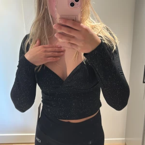 Svart glittrig topp från Nelly - Supersnygg svart topp från Nelly med glittriga detaljer och lång ärm. Toppen har en croppad passform och markerad byst med fyrkantig urringning. Perfekt för dig som vill sticka ut med lite extra glitter.