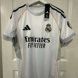 Real Madrid vit matchtröja 2025/26 - Säljer en officiell Real Madrid fotbollströja från Adidas i vitt med svarta detaljer och gula linjer. Tröjan har klubbmärke på bröstet, Adidas-logga och sponsortryck. Tillverkad i lätt och ventilerande material, perfekt för match eller träning.