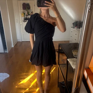 Svart one shoulder kort klänning - Säljer en svart kort klänning med one shoulder och puffärm. Klänningen har en figurnära midja och ett flowy kjolparti. 