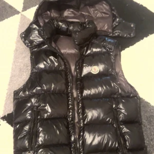 Moncler väst  - Säljer en svart, glansig dunväst från Moncler med huva och dragkedja framtill. Västen har två fickor med dragkedja och klassisk Moncler-logga på bröstet. Perfekt för lager-på-lager och riktigt snygg till streetwear-stilen.