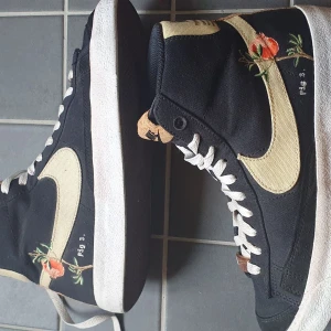 Nike Blazer Mid med broderi, svart/beige - Unika Nike Blazer Mid sneakers i svart canvas med beige swoosh och detaljer i kork. Broderade blommor och texten 'Fig 3.' på sidan ger en kreativ vibe. Vita snören och platt sula. Perfekta för dig som vill sticka ut med stilrena men ändå lekfulla skor. Skicka 9/10 