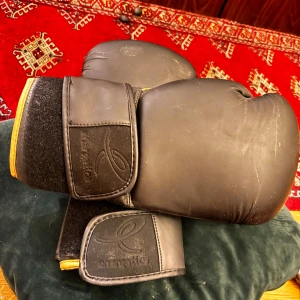 Svarta boxningshandskar Energetics TN - Säljer ett par svarta boxningshandskar från Energetics TN Pro Line, 14 oz. Handskarna har gul text och detaljer, bred kardborrestängning och är tillverkade i skinn. Perfekta för dig som vill träna boxning eller kampsport.