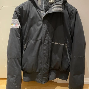 Svart dunjacka Sail Racing Large - Grym svart retro dunjacka från Sail Racing! Logga på bröstet och patch på ärmen. Jackan har huva, dragkedja framtill och två fickor med dragkedja. 
