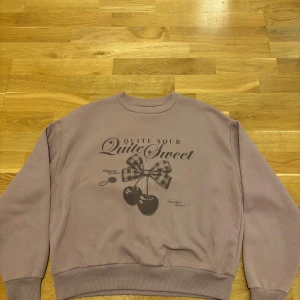 Ljuslila sweatshirt med körsbärstryck - Säljer en ljuslila sweatshirt med tryck av körsbär och rosett framtill samt texten 'Quite Sour Quite Sweet'. Tröjan har rund halsringning, ribbade muddar och en relaxed passform. Perfekt för dig som gillar söta detaljer och pastellfärger.
