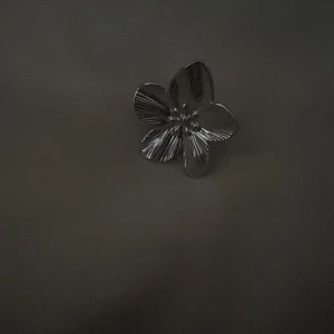 Silverfärgad blomring - Snygg ring i silverfärgat material med en stor blomma som har fem detaljerade kronblad. Blommans yta är lätt räfflad för extra effekt och ger en cool, modern vibe. Perfekt för dig som vill sticka ut med ett unikt smycke.