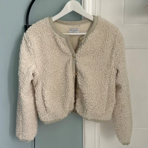 Beige vit teddykofta / jacka i pile - Mysig beige vit teddykofta / jacka i pile från Holly & Whyte by Lindex. Koftan har rund hals, långa ärmar och knäppning framtill med ljusa knappar. 