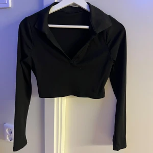 Svart croppad långärmad polotopp - Snygg svart croppad polotopp med lång ärm och djup v-ringning. Toppen har en klassisk krage och är tillverkad i ett stretchigt, ribbat material som sitter tight. Perfekt att styla med höga jeans eller kjol.