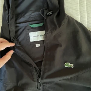 Svart vindjacka från Lacoste med huva - Svart vindjacka från Lacoste med huva och dragkedja framtill. Jackan har vita Lacoste-loggor längs ärmarna och klassisk krokodil på bröstet. Tillverkad i polyester och har två fickor med dragkedja. Perfekt för dig som gillar sportig och stilren look.