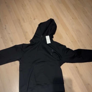 Svart hoodie från Ami Paris - Svart hoodie från Ami Paris med klassisk huva och snörning. Tröjan har en stor magficka och diskret Ami-logga på bröstet. Perfekt för chill dagar och streetwear-stil. Materialet är mjuk bomull och passformen är normal.