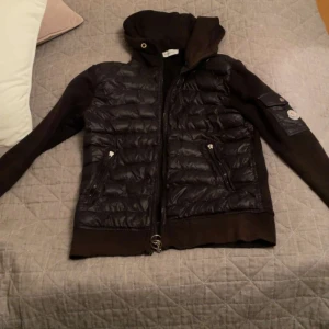 Svart Moncler cardigan  - Svart pufferjacka från Moncler med quiltad front och ärmficka med logga. Jackan har huva, dragkedja med ringdetalj och två fickor framtill. Materialmix med glansig polyester och stickade partier på ärmar och sidor. Perfekt för dig som vill ha en snygg och sportig look.OBS ÄR LITE SÖNDER PÅ ARMARNA 