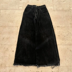 Svarta wide jeans från JNCO - Säljer ett par svarta wide jeans från JNCO med kontrastsömmar och råa, fransiga benslut. Baktill finns stora fickor med blå JNCO-logga i graffiti-stil. Jeansen har hög midja och är tillverkade i kraftigt denim. Perfekta för dig som gillar streetwear och retrovibe.