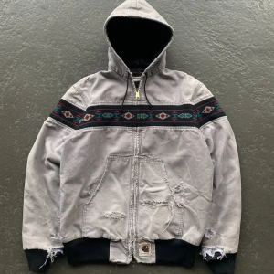 Grå Carhartt hoodiejacka med mönster - Säljer en grå hoodiejacka från Carhartt med färgglada aztekinspirerade mönsterband över bröst och rygg. Jackan har huva, dragkedja och muddar i svart. Praktiska fickor framtill och slitna detaljer för en cool, avslappnad look.