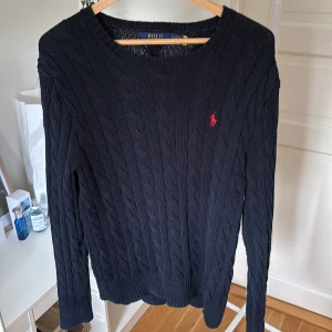 Mörkblå kabelstickad tröja Polo Ralph Lauren - Kabelstickad mörkblå tröja från Polo Ralph Lauren med klassisk röd broderad logga på bröstet. Tröjan har rund halsringning, långa ärmar och ribbade muddar. Perfekt för dig som gillar en tidlös och clean stil. Köpt för 2095kr storlek small