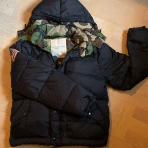 Svart pufferjacka från Ralph Lauren - Säljer en svart pufferjacka från Denim & Supply Ralph Lauren. Jackan har camouflagemönstrat foder , patch med amerikansk flagga på ärmen. Jackan är väldigt unik. Tveka inte med att höra av dig vid frågor /Alex😀🤝