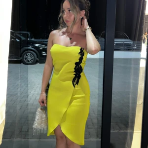 Gul omlottklänning med svart applikation - Bara använd en gång!Säljer en snygg gul kortklänning med omlott-design och strapless modell. Klänningen har en svart dekorativ applikation längs framsidan som ger en cool kontrast. Passformen är tight och materialet känns slätt och festligt. Perfekt för dig som vill sticka ut med färg och detaljer.