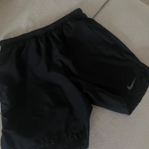 Svarta träningsshorts från Nike - Svarta shorts från Nike med elastisk midja och klassisk logga på benet. Perfekta för träning eller sport, tillverkade i lätt och syntetiskt material som andas. Enkel och stilren design som passar till alla sportiga outfits.