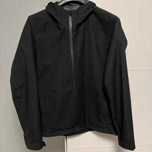 Svart vindjacka från Uniqlo med huva - Svart vindjacka från Uniqlo med minimalistisk design och huva. Jackan har hel dragkedja framtill och är tillverkad i ett vattenavvisande och andningsbart syntetmaterial. Perfekt för blåsiga dagar och enkel att matcha med olika outfits.