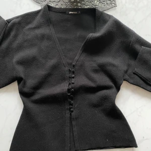 Svart kofta med knappar från Gina Tricot - Säljer en svart kofta från Gina Tricot i storlek S. Koftan har v-ringning, långa ärmar och dekorativa knappar framtill. Materialet är mjukt och stickat, perfekt för lager-på-lager och snygg till jeans eller kjol. De enda bandet har gått av, men går lätt att sy igen 