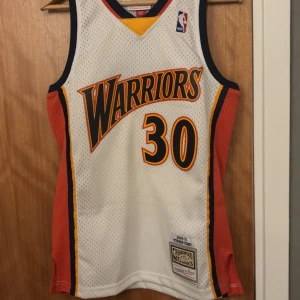 Harwood classic jersey Stephen Curry  - Golden State Warriors baskettröja från Mitchell & Ness, Hardwood Classics 2009-10 Stephen Curry. Vit mesh med marinblå, orange och gula detaljer, stora tryckta siffror och namn. Ärmlös modell med klassisk sportig look och autentiska NBA-loggor.