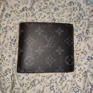 Svart Louis Vuitton plånbok i skinn - Säljer en svart plånbok från Louis Vuitton med klassiskt monogrammönster. Plånboken har flera kortfack, fönster för ID och är tillverkad i exklusivt skinn. Perfekt för dig som vill ha en stilren och lyxig accessoar i fickan.