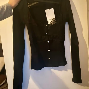 Svart spetsblus från Zara - Supersnygg svart blus från Zara med långa ärmar och vita knappar framtill. Blusen har v-ringning och fina spetsdetaljer vid bysten och över bröstet. Perfekt att styla med jeans eller kjol för en trendig look.