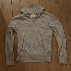 Grå half zip tröja från GANT - Säljer en stilren grå långärmad tröja från GANT i storlek S. Tröjan har en klassisk half zip-dragkedja vid kragen och är tillverkad i 100% bomull. Perfekt för lager-på-lager och enkel att matcha med jeans eller chinos.