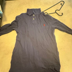 Marinblå pikétröja från Polo Ralph Lauren - Säljer en marinblå långärmad pikétröja från Polo Ralph Lauren i slim fit. Tröjan har klassisk krage, knäppning med två knappar och röd broderad logga på bröstet. Tillverkad i bomull och passar perfekt för en clean och stilren look.
