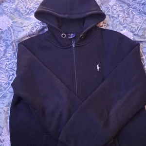 Svart hoodie från Polo Ralph Lauren - Svart hoodie från Polo Ralph Lauren med klassisk vit broderad logga på bröstet. Tröjan har dragkedja framtill, huva med snörningshål och är tillverkad i mjuk bomull. Perfekt för dig som gillar stilren och enkel streetwear.