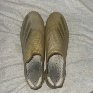 Guldiga adidas fotbollsskor utan snörning - Säljer ett par guldiga adidas fotbollsskor med låg profil och utan snörning. Skorna har en strömlinjeformad design, mönstrad ovandel och fasta dobbar för gräsplan. Tillverkade i syntetmaterial och perfekta för dig som vill sticka ut på planen. Högsta moddelen köpte för 3000kr