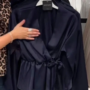 Marinblå omlottblus från Missäy - Snygg marinblå omlottblus från Missäy i glansigt satinmaterial. Blusen har långa ärmar, v-ringad hals och knyt i midjan. Perfekt att styla med jeans eller kjol.