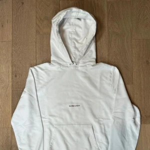 Vit hoodie från Saint Laurent - Snygg vit hoodie från Saint Laurent med minimalistisk logga på bröstet. Klassisk känguruficka, huva med snörning och ribbade muddar. Perfekt för dig som gillar stilren och clean streetwear.