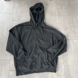 Svart hoodie med dragkedja från H&M - Svart hoodie från H&M i relaxed fit med dragkedja framtill och två fickor. Tröjan har huva med snörning och är tillverkad i mjuk bomullsmix. Perfekt för chill dagar eller när du vill ha en enkel och stilren look.