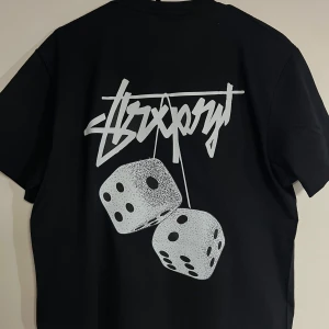 Svart Stüssy t-shirt med tärningar - Svart t-shirt från Stüssy med Dover Street Market London-tryck fram och stort grafiskt tryck med tärningar och logga på ryggen. Klassisk rund hals och korta ärmar. Skön passform och tillverkad i mjuk bomull, perfekt för streetwear-stilen.