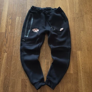 Svarta Nike mjukisbyxor med tryck - Svarta mjukisbyxor från Nike med rosa logga och coolt tryck på ena benet. Byxorna har dragsko i midjan, ribbade muddar vid bensluten och en lång dragkedja längs ena fickan. Perfekta för chill eller träning.