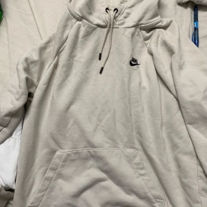 Beige Nike hoodie med broderad logo - Snygg beige hoodie från Nike med broderad svart logga på bröstet. Klassisk känguruficka framtill, snörning i huvan och mjukt bomullsmaterial. Perfekt för chill dagar eller när du vill ha en clean och enkel look.