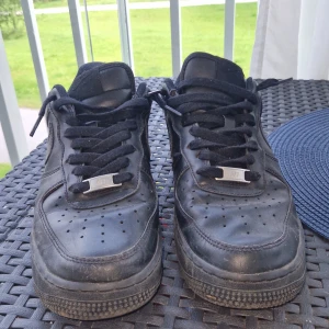 Svarta Nike Air Force 1 sneakers - Klassiska helsvarta Nike Air Force 1 sneakers i skinn med svarta snören och perforerad tå. Snygg och tidlös modell med låg profil och robust sula. Perfekta för dig som gillar streetstyle och vill ha en ikonisk sneaker i garderoben.