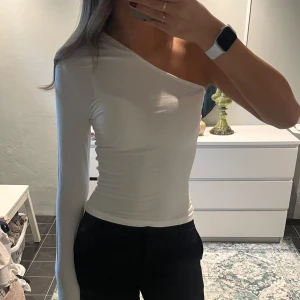 Vit oneshoulder topp med lång ärm - Säljer en stilren vit oneshoulder topp med en lång ärm och tight passform. Toppen är enkel men ändå unik med sin asymmetriska design och passar perfekt till svarta byxor eller kjol. Materialet känns mjukt och stretchigt, vilket gör den bekväm att bära.
