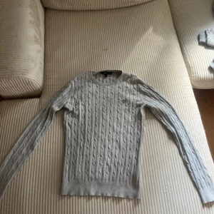 Ljusgrå kabelstickad tröja från GANT - Snygg ljusgrå kabelstickad tröja från GANT med rund hals och diskret broderad logga på bröstet. Tröjan har lång ärm och är i ett mjukt bomullsmaterial, perfekt för lager-på-lager under kyligare dagar.