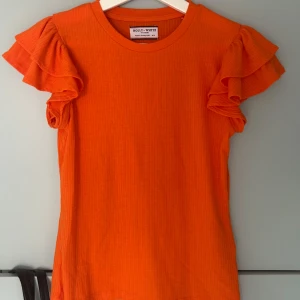 Orange volangtopp  - Snygg orange topp från Holly & Whyte (Lindex) med ribbad struktur och volangärmar. Rund halsringning och figurnära passform. Skönt mjukt tyg.