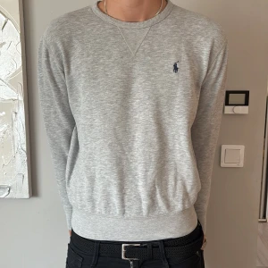Grå sweatshirt från Polo Ralph Lauren - Snygg grå sweatshirt från Polo Ralph Lauren med klassisk rund hals och broderad marinblå logga på bröstet. Tröjan har ribbade muddar vid ärmslut och nederkant, samt en diskret V-detalj vid halsen. Perfekt för chill dagar och enkel att matcha med jeans.