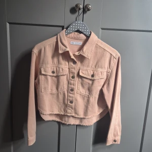 Beige croppad jeansjacka från Zara - Snygg croppad jeansjacka från Zara i beige med rå kant nertill. Jackan har två bröstfickor med knappar, klassisk krage och långa ärmar. Perfekt för dig som vill ha en chill och trendig look. Passar till det mesta och är lätt att matcha.