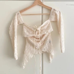 Vit omlottopp med snörning och volang - Supersöt vit omlottopp med långa ärmar och transparent vågig struktur. Toppen har snörning framtill och volangdetaljer längs kanterna. Den är croppad med spetsig nederkant och ger en romantisk vibe. Perfekt för dig som gillar unika och trendiga plagg!