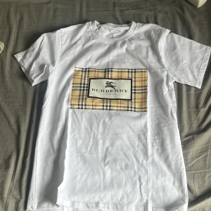 Vit Burberry t-shirt med rutig patch - Vit t-shirt från Burberry i storlek S med klassisk rutig patch framtill och Burberry-logga. T-shirten har rund hals och korta ärmar. Materialet är mjuk bomull och passformen är normal. Perfekt för dig som gillar stilrena och ikoniska designer.