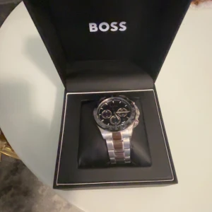 Stilren silverfärgad BOSS klocka - Snygg klocka från BOSS med svart urtavla och detaljer i guld. Klockan har en robust länk i silverfärgat metall och flera små urtavlor för extra funktioner. Perfekt accessoar för dig som vill ha en klassisk men modern look. Den finns i gbg kvitto finns med 