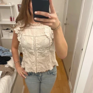 Vit blus med volang och spetsdetaljer - Supersöt vit blus med volangdetaljer på axlar och nederkant. Blusen har fina spetsinslag och broderade partier framtill, samt en liten knappöppning i nacken. Perfekt till jeans för en fräsch look.