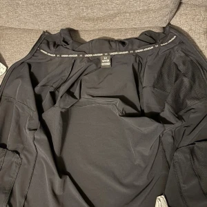 Svart vindjacka från Under Armour - Svart vindjacka från Under Armour med huva och dragkedja hela vägen. Jackan har en bröstficka med dragkedja, meshpaneler på insidan och reflekterande detaljer på axlarna. Perfekt för dig som gillar sportig stil och vill ha något lätt och smidigt.