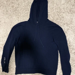 Mörkblå hoodie i kashmir från RLX - Mörkblå hoodie från RLX Ralph Lauren i mjuk tvättbar kashmir. Tröjan har huva med snörning, långa ärmar och coola elbow patches i mörkare tyg. RLX-logga broderad på ena ärmen. Perfekt för dig som gillar stilren och lyxig.