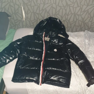 Svart pufferjacka från Moncler - Säljer en svart, glansig pufferjacka från Moncler med huva och dragkedja med rödvit detalj. Jackan har en bröstficka med Moncler-logga och stora fickor framtill. Perfekt för kalla dagar och riktigt snygg streetstil.